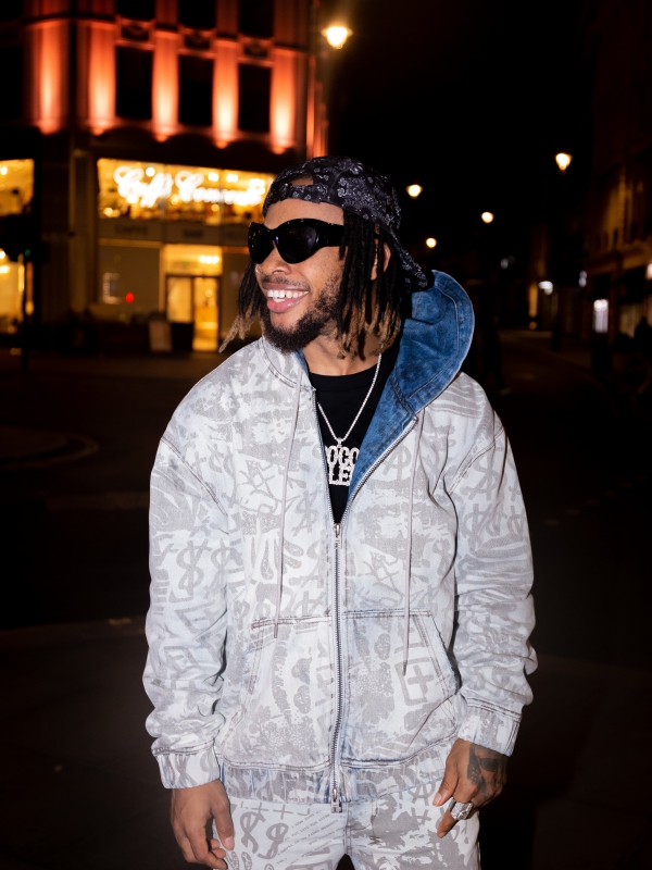 A Man In A Patterned Grey Tracksuit And Beanie Smiling On A City Street At Night.